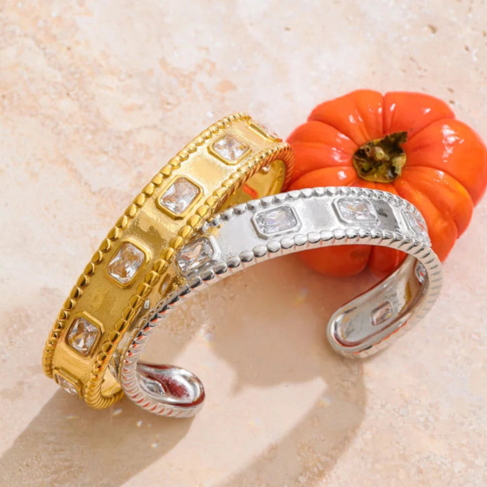 Two gold and silver bracelets on a textured beige surface with a small orange pumpkin.