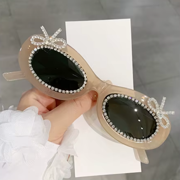 Beige sunglasses with decorative elements held by a hand on a gray surface.