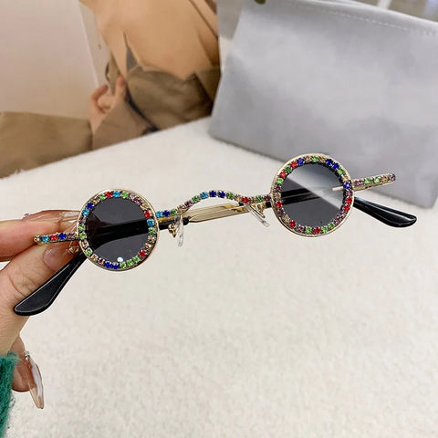 Round sunglasses with colorful gemstones held by a hand on a white surface.